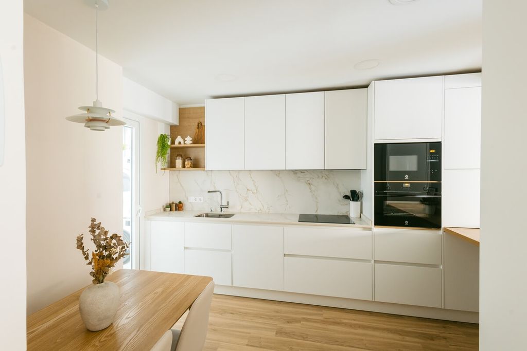 En este caso optamos por frentes blancos mate, una elección que potencia la luminosidad y sensación de amplitud del espacio. Los acabados blancos siguen siendo una tendencia atemporal en el diseño de cocinas, especialmente cuando se combinan con detalles en madera como en este proyecto, donde el suelo y la encimera del desayunador aportan la calidez necesaria.